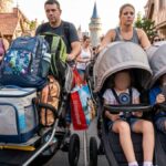 Parents pushing massive double strollers overloaded with heavy backpacks, diaper bags, coolers, and souvenir bags down a crowded path in Fantasyland at Disney World.