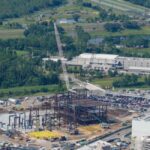 An aerial landscape view of a massive theme park construction site showing a large steel framework building rising into the air with a tall white crane next to a parking lot and background highway.
