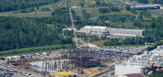An aerial landscape view of a massive theme park construction site showing a large steel framework building rising into the air with a tall white crane next to a parking lot and background highway.