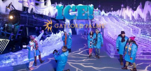 Visitors wearing blue parkas posing in front of a giant ice sculpture train with "ICE! Gaylord Palms" branding overlay.