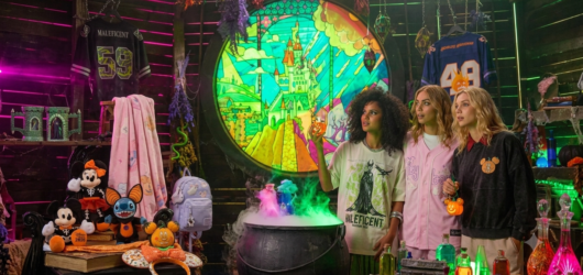 Three women modeling Disney Halloween apparel including a Maleficent graphic tee, pastel pink baseball jersey, and Mickey pumpkin sweatshirt in a spooky potion lab setting filled with plush toys and holiday accessories.