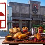 A featured image for a news article summarizing Six Ravens at Disney Springs. The composition features a large, detailed red logo of the crowned raven profile on the left. The background is a wide-angle photograph of the tavern exterior with its primary shield logo. In the foreground, a detailed rustic wooden table holds a full meal: multiple savory Coffyn hand pies, buckets of Tale Teller Tatties smashed potatoes, various dipping sauces, a metal mead tankard, and a pint glass of amber craft beer.