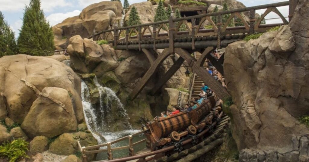 Seven Dwarfs Mine Train: Magic Kingdom Ride Guide 2 Seven Dwarfs Mine Train roller coaster vehicle curving past a waterfall and rock bridge in Fantasyland at Magic Kingdom.