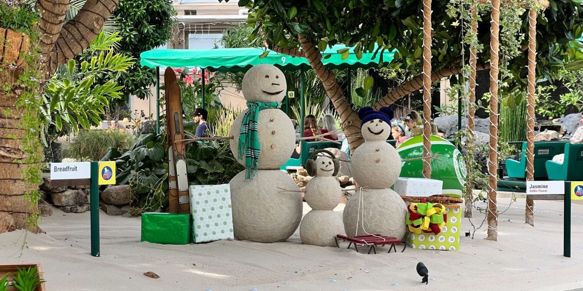 Sand snowman family with winter scarves, skis, and gift boxes as a green Living with the Land boat passes behind.