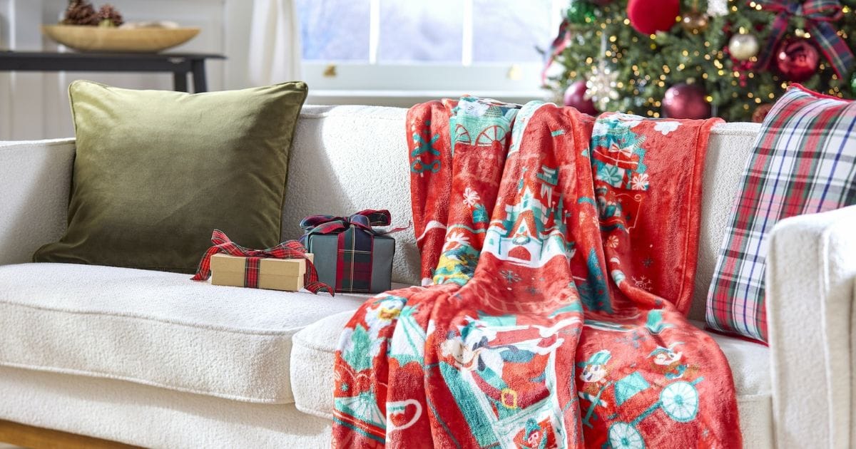 Red Disney holiday fleece throw blanket draped over a living room sofa next to plaid pillows and wrapped gifts.