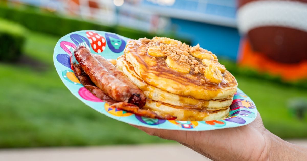 Decadent stack of hot pancakes topped with caramelized bananas and crumb streusel alongside breakfast sausage links and bacon