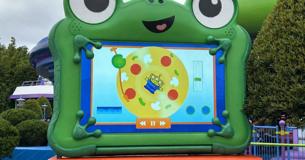 The green frog tablet character Lilypad displays an interactive screen showing a Pizza Planet Alien centered on a spinning pizza record with playback controls, located at Disney's Hollywood Studios