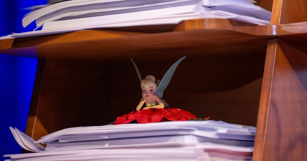 Small Tinker Bell figure resting on a red poinsettia petal tucked between stacks of animation paper on a wooden studio shelf