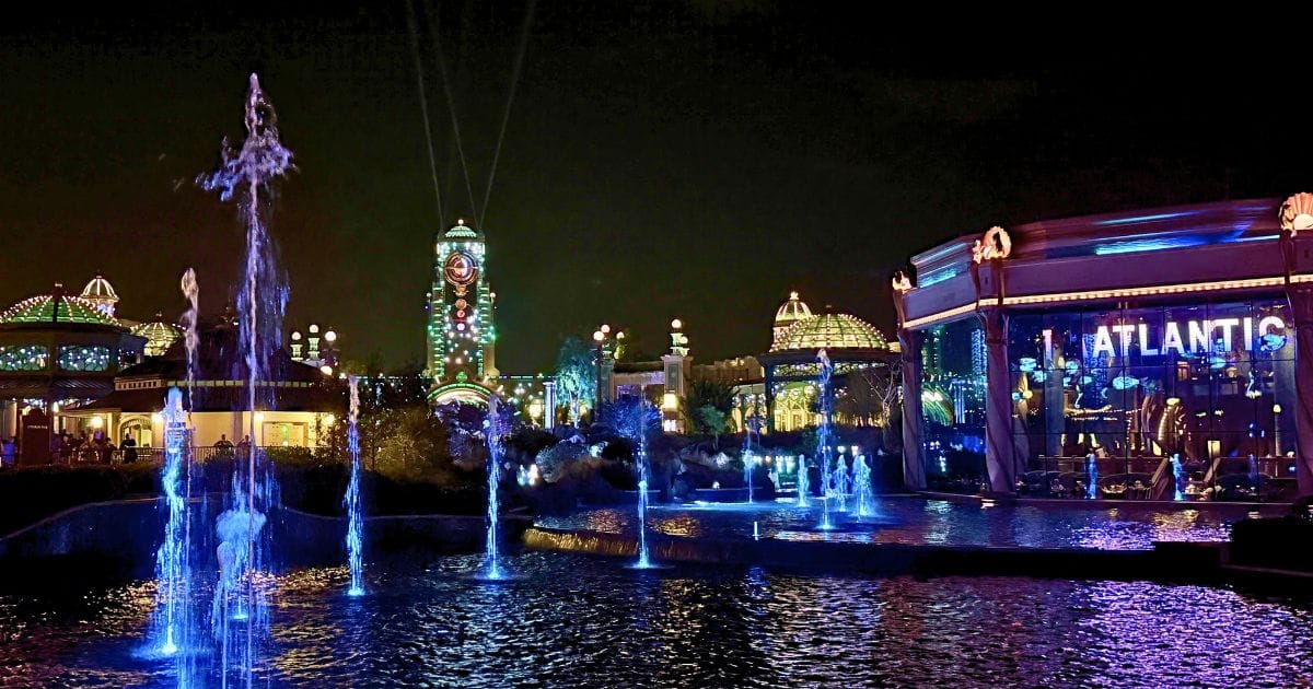 Illuminated blue fountains dancing in front of The Atlantic restaurant in Celestial Park during an Epic Universe night event.